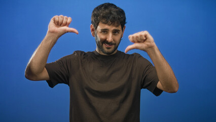 Fototapeta premium Man points thumbs to chest in studio against blue backdrop, bare thumbs and forearms visible, beard and slight smile while facing camera; confidence.