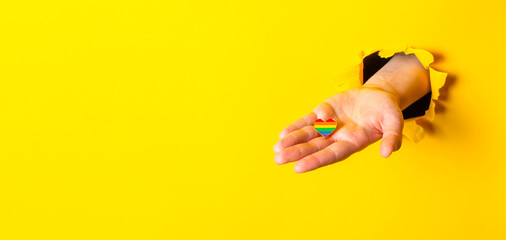 Fototapeta premium Hand Holding Rainbow Heart Through Yellow Paper Background
