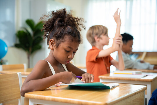 Diligent african american schoolgirl writing at a desk while a classmate raises his hand in classroom. Diverse group of multiethnic primary students participating in a lesson under active learning.