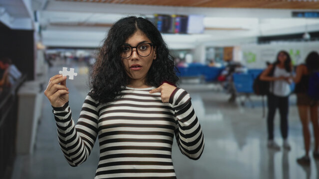 Woman holding a puzzle piece and pointing finger to chest in an airport terminal, striped shirt visible; confusion uncertainty travel stress.