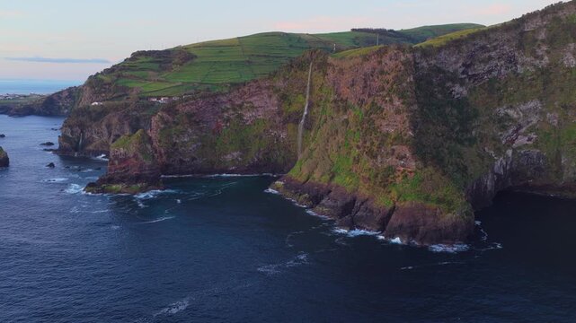 Aerial view of the dramatic coastal cliffs and waterfall at Miradouro dos Caimbros, featuring lush green fields above the Atlantic Ocean in Cedros, Acores, Portugal.