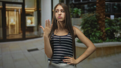 Fototapeta premium Teenage girl with long hair holding hand near mouth in a dismissive pout gesture inside a building, palm raised and one hand on hip; indifference attitude.