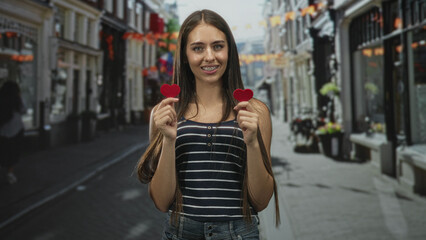 Fototapeta premium Teenage girl holding two red felt hearts in hands on a street, smiling with braces and slight head tilt while presenting the hearts; love romance joy.