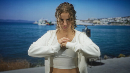 Fototapeta premium Woman with visible navel piercing clasping hands at chest beside a harbor, wearing white crop top and zip hoodie, curly hair framing face and midriff exposed; confidence.