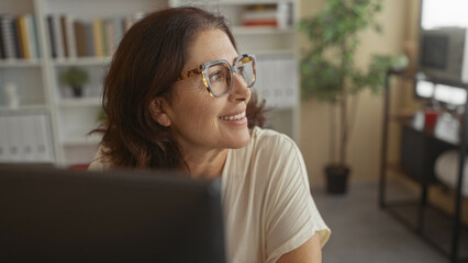 Woman with tortoiseshell glasses smiles toward a computer monitor while seated at a desk in an office building with bookshelves and plant visible  contentment focus. © Krakenimages.com