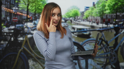 Fototapeta premium Woman with red hair holding a laptop and touching her nose with one finger while standing on a bicycle lined canal street with blurred canal boats; thoughtful remote work.