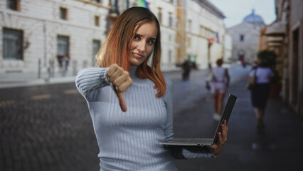 Fototapeta premium Woman holding laptop with thumb down gesture on a cobblestone street beside a historic building, redhead in a light sweater; disapproval critique feedback.