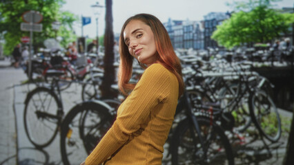 Naklejka premium Woman posing with bicycle, smiling and turning head on a street lined with parked bikes; quiet confidence.