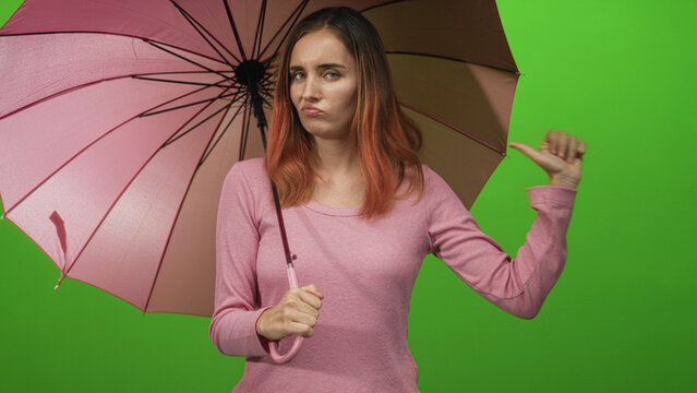 Young woman with red hair holding a pink umbrella and gesturing thumb to the side while wearing a pink sweater in a studio; displeasure.