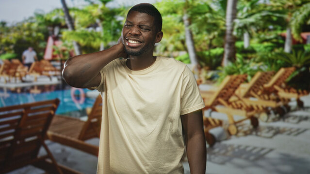 Man hand on neck wincing with neckache against a studio pool backdrop with loungechairs and palms; vacation discomfort.
