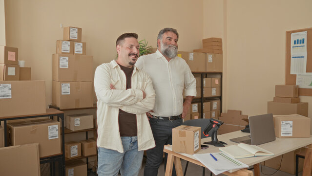 Two men standing with arms crossed beside cardboard packages in a packing building, smiling and checking inventory; teamwork pride.