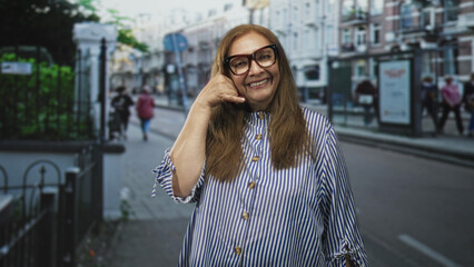 Fototapeta premium Woman with large glasses and striped shirt making a phone hand gesture on a city street; warm connection joy.