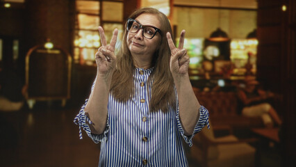 Fototapeta premium Woman holding up two peace signs with both hands in a hotel building interior; playful warm confidence.