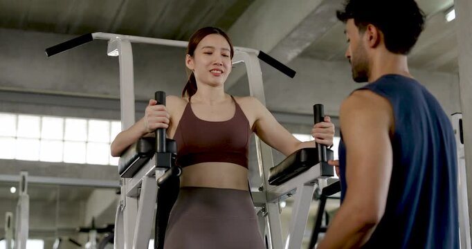 Young Asian couple smiling during gym coaching session, woman stands on knee raise machine while man gives supportive guidance, partner workout, leg training, motivation, wellness lifestyle.