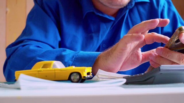 Car insurance agent calculating cost with calculator and smartphone close up. Focus on hands, yellow toy car on desk. Shallow depth of field, natural light. Businessman estimating vehicle policy.