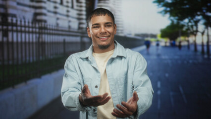 Obraz premium Man smiling, hands open and gesturing while standing on a street beside a stone wall and iron fence in front of a building; optimism approachability.