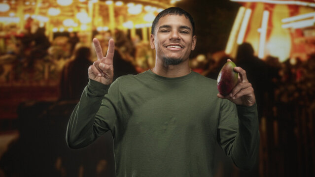 Man holding mango and flashing peace sign on street market with warm lights and blurred crowd; joyful celebration.