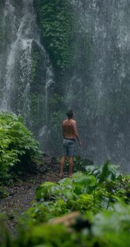 Man with raised arms under powerful waterfall, feeling energy in tropical jungle nature