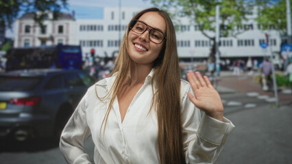 Fototapeta premium Woman smiling and waving hand on a busy street in front of shops and cars, wearing glasses and white blouse; friendly greeting.