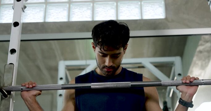 Young Asian man pauses at barbell rack during shoulder workout in modern gym, gripping weight bar and looking down between sets, solo strength training, recovery, wellness, healthy lifestyle.
