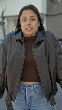 Young woman in brown top and leather jacket shrugs shoulders, showing midriff and blue jeans beside parked car on street; bemusement.