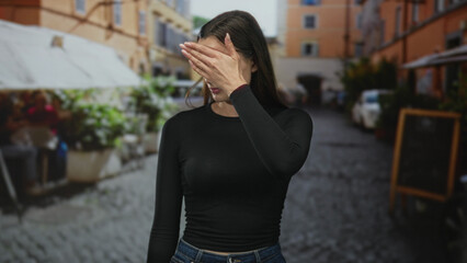 Fototapeta premium Young caucasian woman holds hand up palm out in front of face on cobblestone street by outdoor cafe, eyes closed and grimacing; defiance boundary.