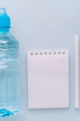 Water bottle, blank notepad and pen for hydration tracking on blue background. © Anastassiya 