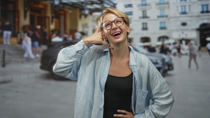 Fototapeta premium Woman making call gesture with hand to ear, wearing glasses and light blue overshirt on a busy street with cars and building facades; playful confidence.