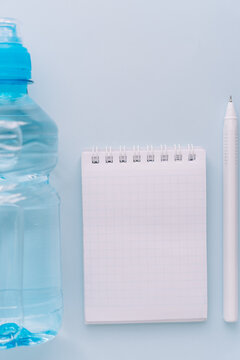 Water bottle, blank notepad and pen for hydration tracking on blue background.