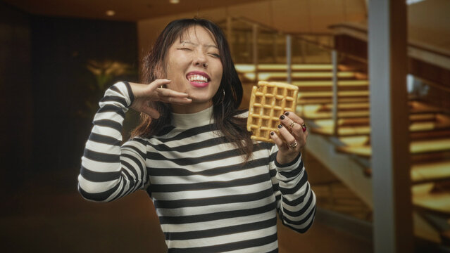 Woman in striped shirt holding a waffle and pointing finger to lip by a lit staircase in building, showing nose ring and hoop earring; playful indulgence.