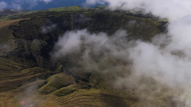 Aerial view of the volcanic crater of Caldeira do Faial with lush green slopes and clouds drifting inside the caldera in Horta, Azores, Portugal.