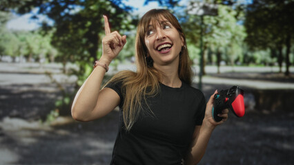 Woman wearing black t shirt holding red and black game controller points index finger up on a forest path smiling broadly  playful gaming moment. © Krakenimages.com