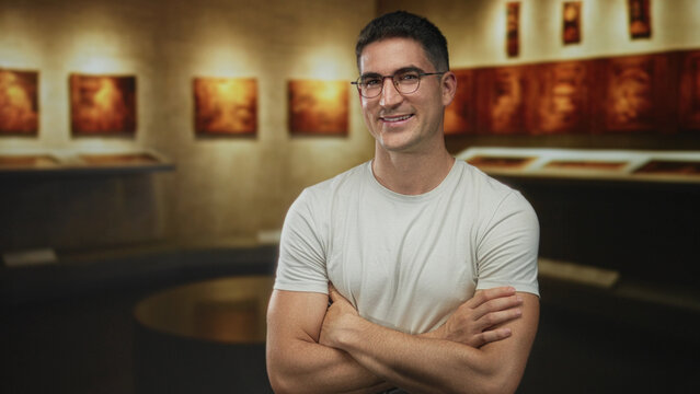 Man with arms crossed, bare forearms visible, wearing white t shirt and glasses in a building art gallery with warm framed paintings and display cases; confidence creativity.