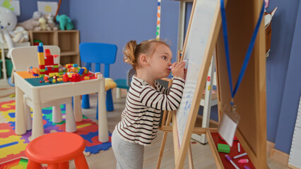Little blonde girl drawing on a whiteboard with a purple marker at a wooden easel in a daycare building, small hand on board and intent expression  curiosity learning play. © Krakenimages.com