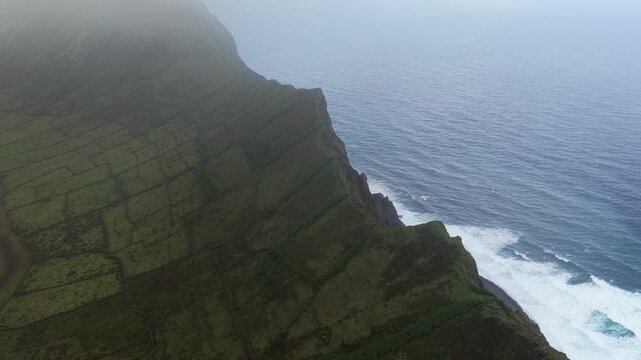 Aerial view of the lush green volcanic crater of Caldeirao with stone walls and steep cliffs overlooking the Atlantic Ocean in Vila do Corvo, Acores, Portugal.