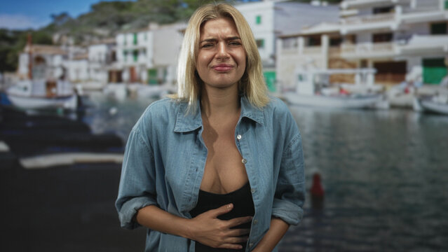 Woman clutching stomach with hand, squinting and grimacing, denim shirt unbuttoned showing cleavage against studio backdrop of a harbor; nausea discomfort.