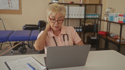 Fototapeta premium Senior woman physiotherapist holding neck while seated at laptop with stethoscope and treatment table in clinic; professional concern.