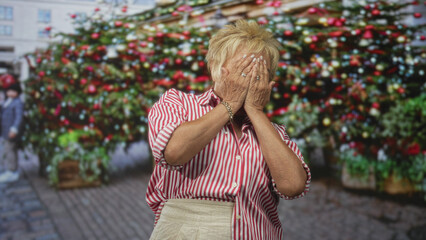 Fototapeta premium Senior hispanic woman covers mouth with hands in street by a large decorated christmas tree and ornaments; surprise nostalgia.