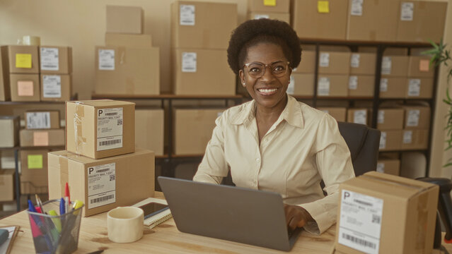 Woman business owner typing on laptop, hands on keyboard among stacked shipping boxes, labels and packing tape on desk in a building; small business confidence.