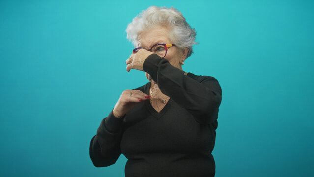 Elderly woman in black sweater pinches her nose with hand and closes eyes in turquoise studio beside glasses; disgust aversion.