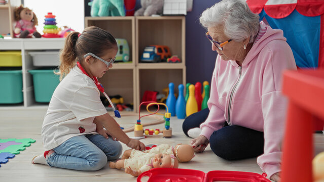 Woman kneeling with girl roleplaying doctor, holding a toy stethoscope and examining a baby doll in a daycare playroom; intergenerational bonding tenderness.