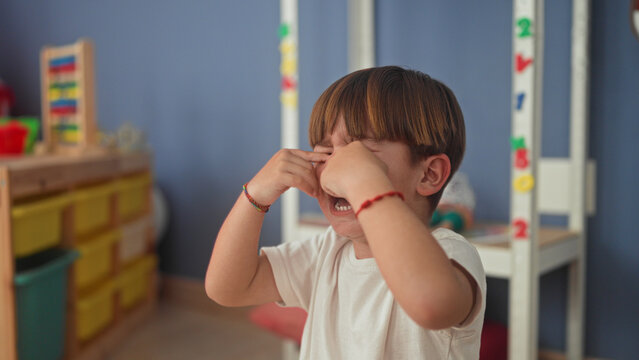 Boy rubbing hands over his eyes and crying with mouth open, visible bracelets and white shirt in a building playroom; sadness comfort.
