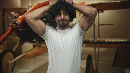 Man with curly hair and beard, hands on head beside vintage biplane in museum exhibit, wearing white tshirt and showing biceps  nostalgia amusement. © Krakenimages.com