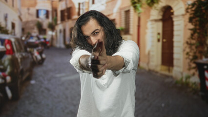 Fototapeta premium Man pointing finger like gun toward camera on old town street with long hair and white shirt; mischief.