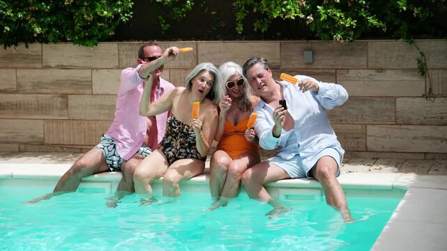Four happy senior friends sitting by the pool, eating popsicles and taking a selfie