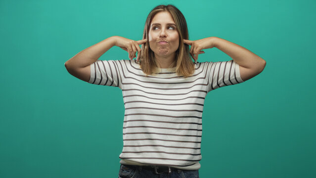 Woman covering ears with hands and rolling eyes, wearing striped white shirt and jeans in studio teal backdrop; annoyance blocking noise.
