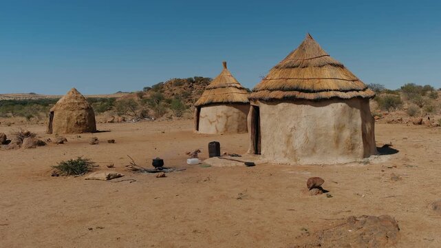 African himba village with mud huts and a cooking pot smoking over a fire in a dry, desert landscape in namibia