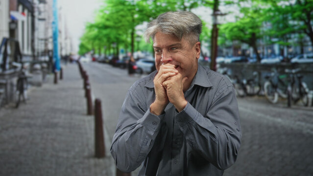Man clasping hands to mouth on a street, biting knuckles with tense posture and visible hands near face; anxiety vulnerability.
