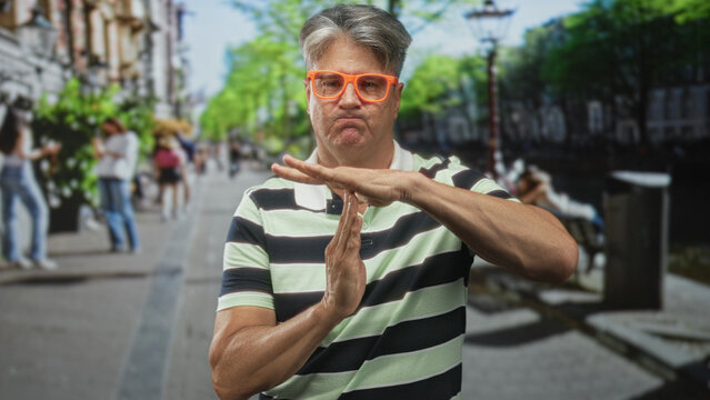 Man with orange glasses making a time out hand gesture on a crowded city street near a canal bench, facing camera; boundary frustration.