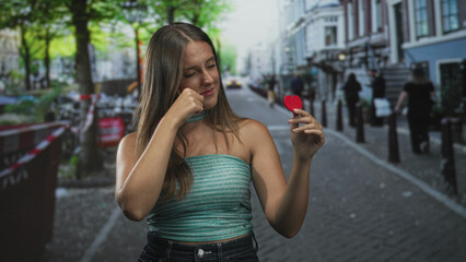 Fototapeta premium Young hispanic brunette woman holding small red felt heart while rubbing her eye on a cobblestone street with outdoor cafes; heartbreak love memory.